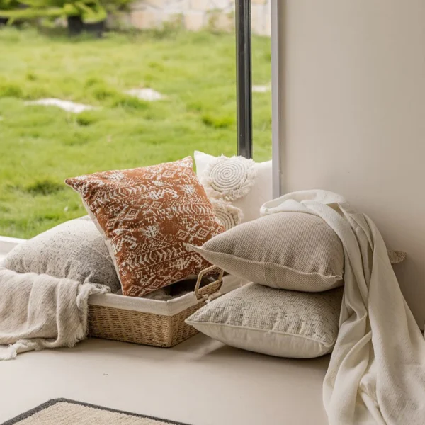 Housse de coussin ethnique berbère Titawin terracotta avec motifs blancs, disposée sur un banc près d'une fenêtre.