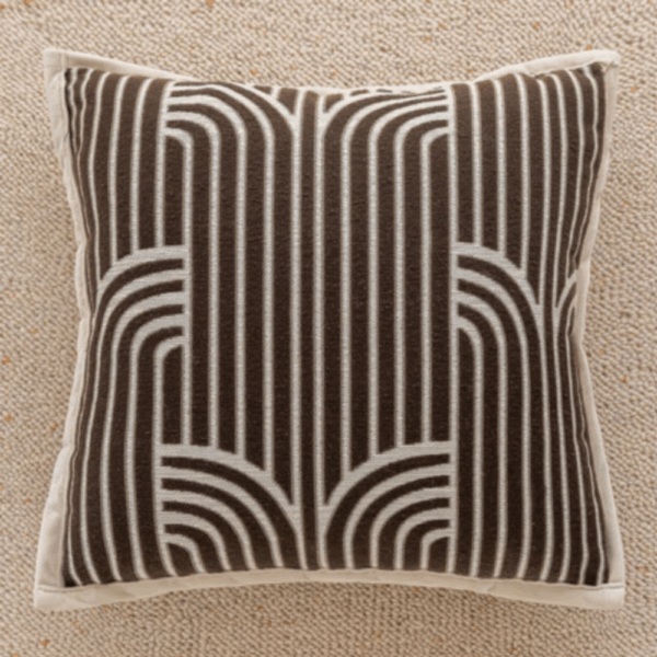 Coussin carré 'Greek' avec un motif géométrique et architectural de style Art Déco, brodé en marron chocolat sur un fond beige, posé sur une surface texturée.